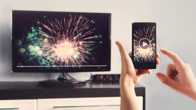 Eine Hand hält ein Smartphone vor einem Smart-TV, auf beiden Geräten ist das gleiche Bild zu sehen, ein Feuerwerk.