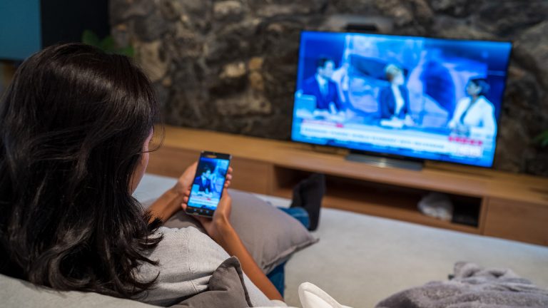 Eine Frau sitzt auf einem Sofa, in der Hand ein Smartphone, dessen Inhalt auch auf dem Fernsehgerät vor ihr wiedergegeben wird.