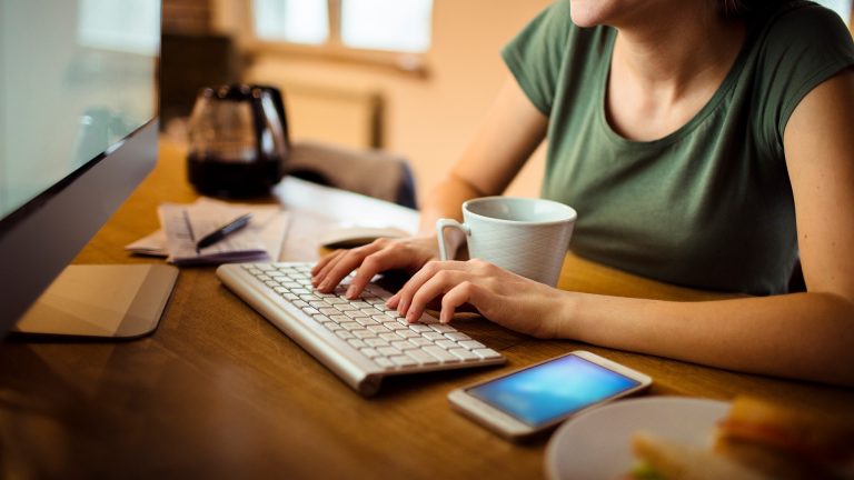 Eine Person sitzt vor einem Monitor und tippt auf einer Tastatur. Vor ihr liegt ein Smartphone.