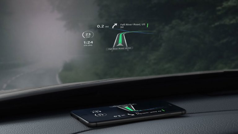 Ein Smartphone liegt auf dem Armaturenbrett eines Autos, direkt unter der Windschutzscheibe. Das Display projiziert den Fahrtverlauf einer Navigationsapp an die Scheibe.