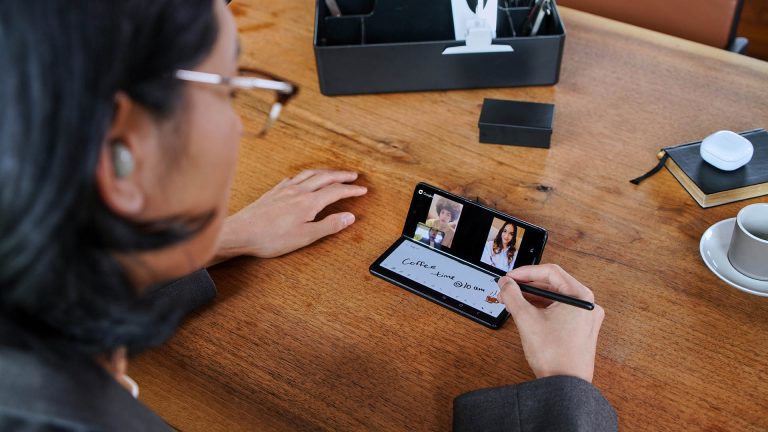 Eine Person sitzt vor einem halb aufgeklappten Samsung Galaxy Z Fold3 5G und schreibt mit einem S Pen auf der unteren Bildschirmhälfte, während die obere ein Videotelefonat zeigt.