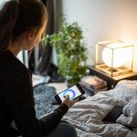 Eine Person sitzt auf einem Bett und steuert per Smartphone eine Lampe vor ihr.