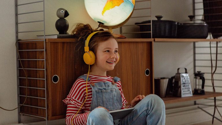 Ein Kind lehnt an einen Schrank. Auf den angewinkelten Knien ruht ein Tablet. Das Kind trägt gelbe Kopfhörer auf den Ohren.
