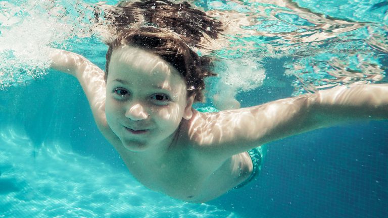 Ein Kind schaut in die Kamera und befindet sich dabei knapp unter der Wasseroberfläche.