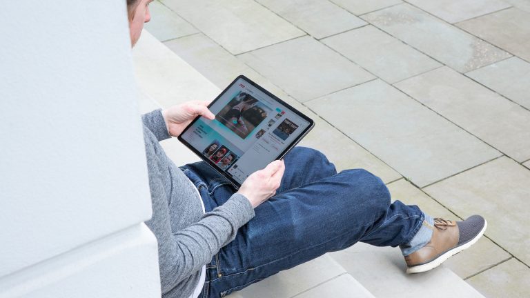 Eine Person sitzt auf einer Treppe an einer Säule gelehnt. In den Händen hält sie ein iPad Pro, auf dem gerade eine Website aufgerufen ist.