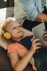 Ein Kind hört über Kopfhörer etwas von einem Tablet. Daneben sitzt ein Elternteil.
