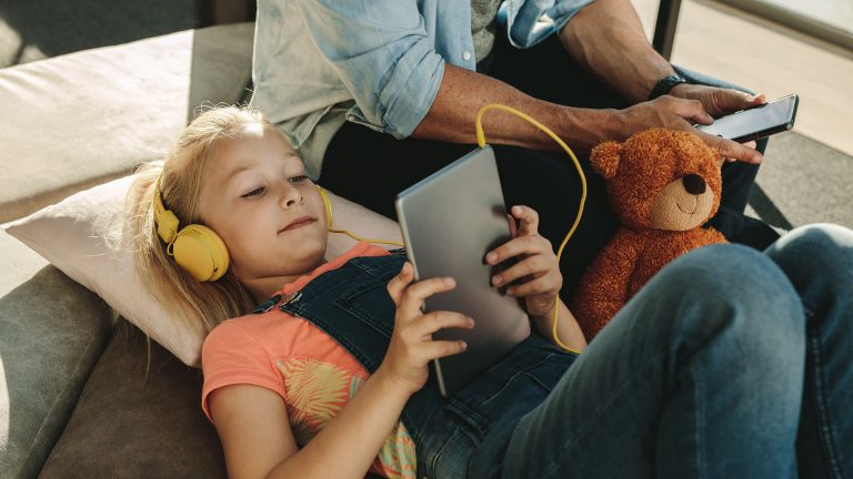 Ein Kind h&ouml;rt &uuml;ber Kopfh&ouml;rer etwas von einem Tablet. Daneben sitzt ein Elternteil.