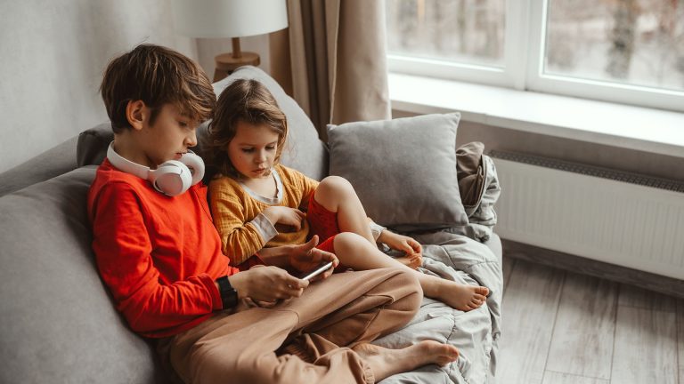 Zwei Kinder sitzen auf einem Sofa und schauen zusammen auf ein Smartphone. Eines der Kinder trägt Kopfhörer.