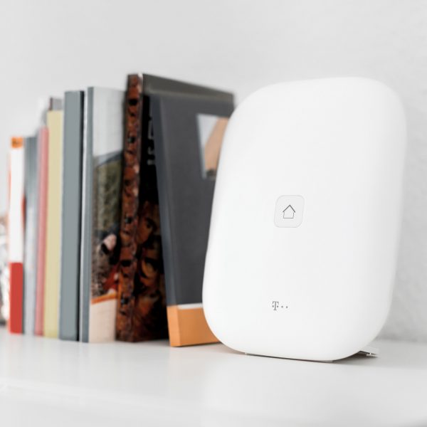 Eine Telekom Magenta Smarthome Home Base steht auf einem Regal neben B&uuml;chern.