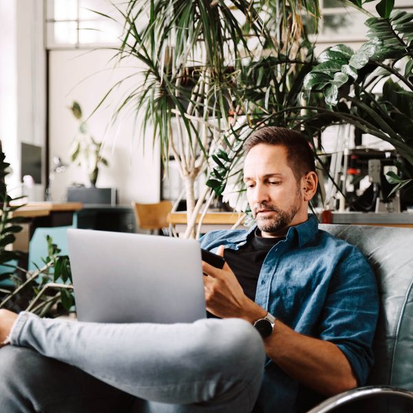 Eine Person sitzt entspannt auf einem Sessel, hat einen Laptop auf dem Scho&szlig; und ein Smartphone in der Hand.