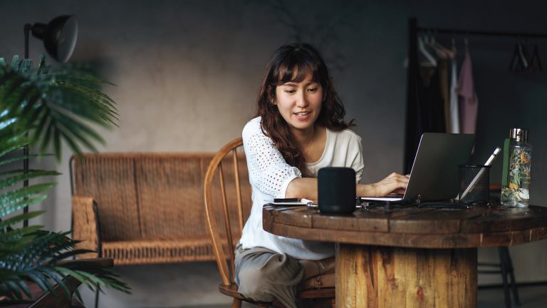 Eine Person sitzt an einem Tisch, vor ihr steht ein aufgeklappter Laptop. Sie spricht zu einem smarten Lautsprecher.