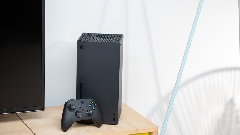 Eine Xbox Box Series X steht auf einem Holztisch neben einem Fernseher.