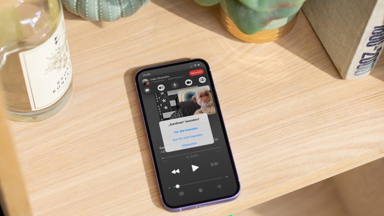 Ein iPhone liegt auf einem Holzregal. Auf dem Bildschirm ist ein Screenshot von Apple Musik zu sehen mit einem eingeblendeten Hinweis von FaceTime SharePlay.