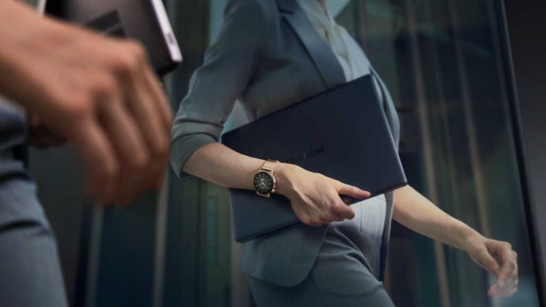 Eine Person mit einem Huawei-Notebook und einem Anzug geht einen Gang entlang. Am Handgelenk trägt sie die Huawei Watch GT 3 in Gold.