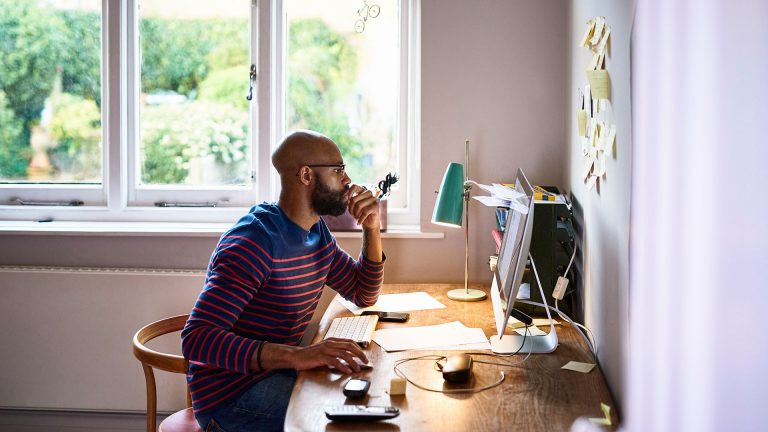 Eine Person sitzt an einem Schreibtisch und schaut auf einen iMac. Neben Maus und Tastatur liegen weitere Arbeitsutensilien auf dem Tisch, unter anderem ein Telefon und mehrere Dokumente.