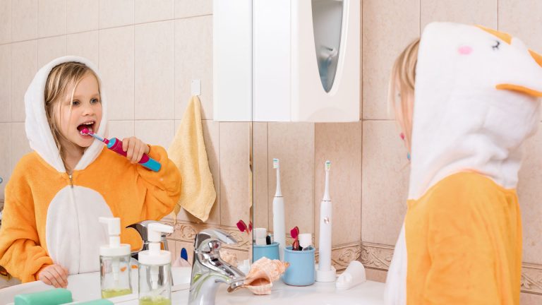 Ein Kind steht vor dem Waschbecken und betrachtet sich beim Zähneputzen mit der elektrischen Zahnbürste im Spiegel.