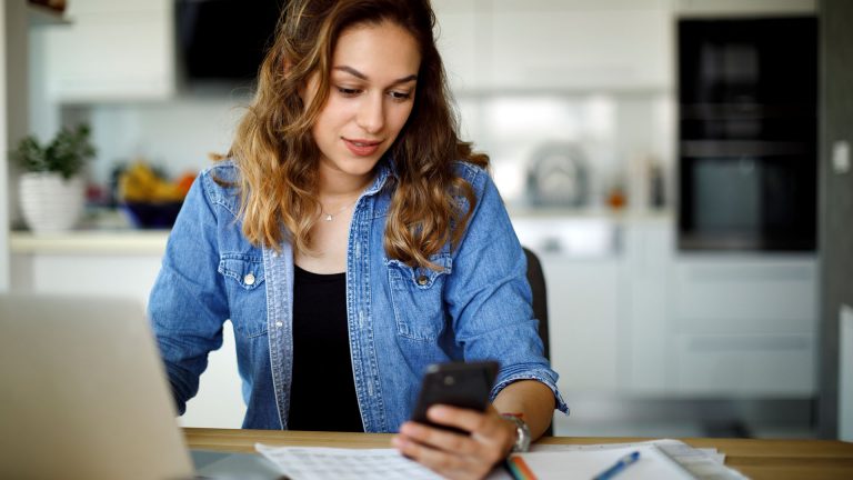 Eine Person sitzt vor einem Laptop und schaut auf ihr Smartphone.