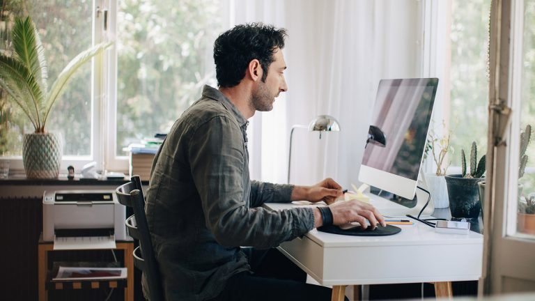 Eine Person sitzt vor einem iMac. Im Hintergrund ist ein Multifunktionsdrucker zu sehen.