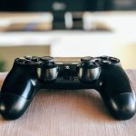 Ein DualShock-4-Controller) liegt auf einer Sofalehne. Im Hintergrund ist ein Fernseher zu erkennen.