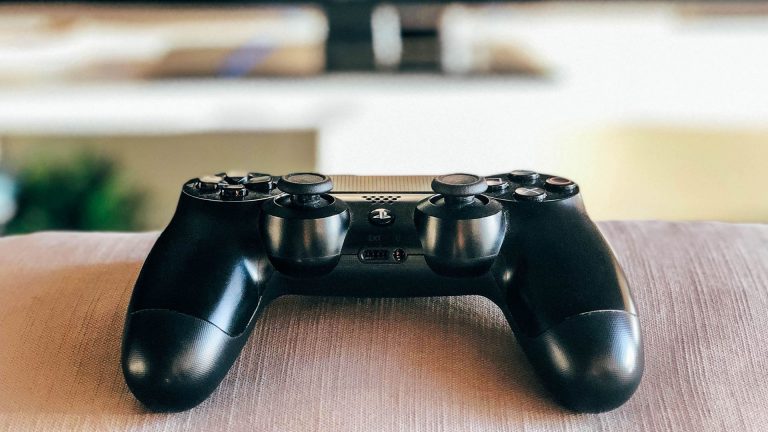 Ein DualShock-4-Controller) liegt auf einer Sofalehne. Im Hintergrund ist ein Fernseher zu erkennen.