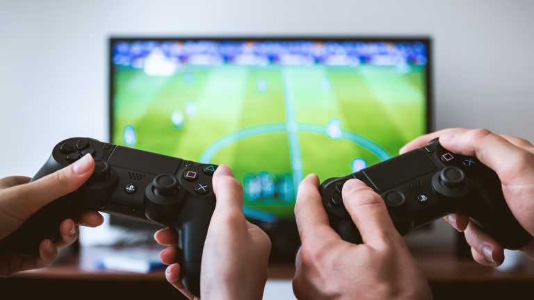 Zwei Paar Hände halten zwei PS4-Controller fest. Im Hintergrund ist ein Fernseher zu erkennen, auf dem ein FIFA-Spiel läuft.