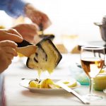 Eine Person schiebt Kartoffeln und K&auml;se mit einem Holzl&ouml;ffel aus einer Raclettepfanne.