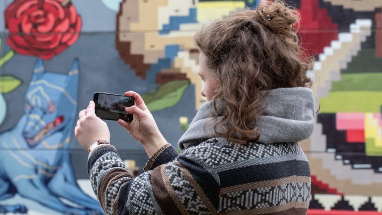 Eine Person hält ein Smartphone in der Hand und fotografiert damit ein Graffiti.