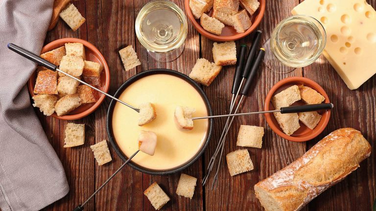 Ein Fonduetopf, gefüllt mit Käse, steht auf einem Holztisch, daneben stehen mehrere Schalen mit in Stücke geschnittenem Brot.