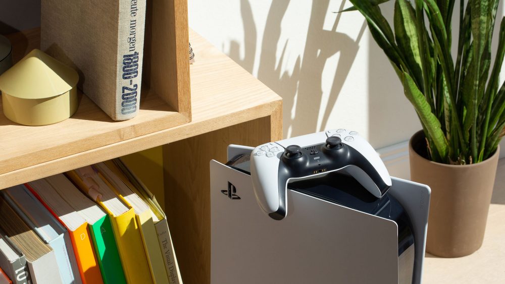 Eine PlayStation 5 steht vor einem Bücherregal. Auf der Konsole liegt ein DualSense-Controller.