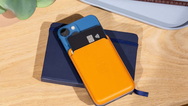 Auf einem Holztisch liegt ein iPhone 12 mini dekorativ auf einem blauen Notizbuch. Die Rückseite des iPhones ziert ein hellbraunes Leder Wallet.