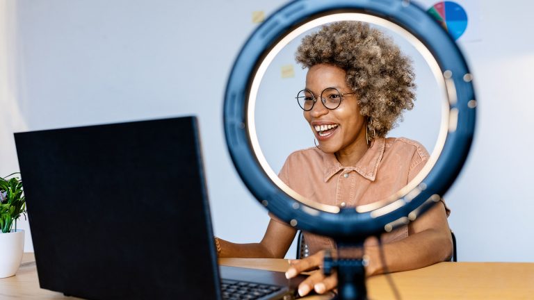 Eine Person sitzt vor einem Laptop. Neben ihr steht ein Ringlicht, das sie anstrahlt.