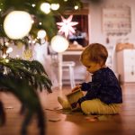 Ein Kleinkind sitzt mit einem Holzpuzzle vor dem Weihnachtsbaum auf dem Boden.
