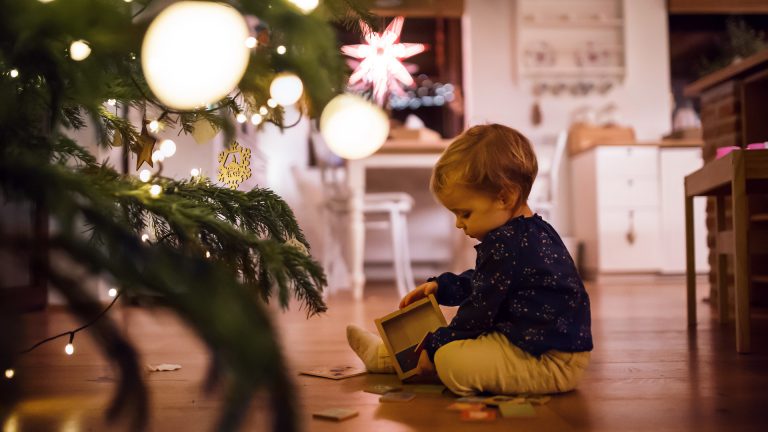 Ein Kleinkind sitzt mit einem Holzpuzzle vor dem Weihnachtsbaum auf dem Boden.