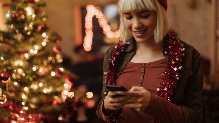 Eine junge Frau steht mit Ihrem Smartphone vor einem mit Lichterketten geschmückten Weihnachtsbaum.