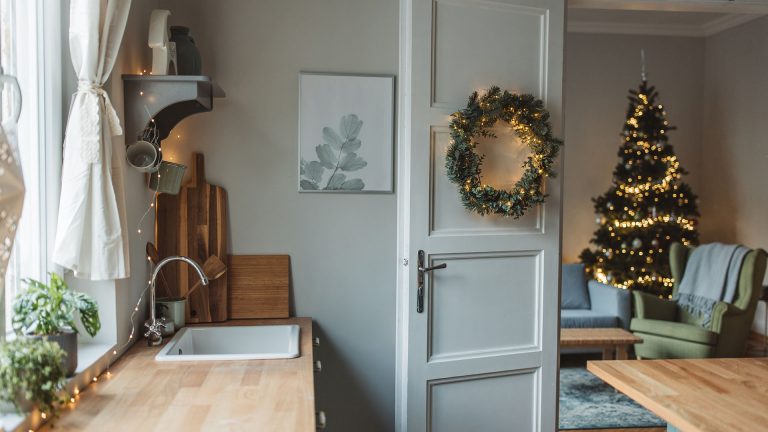 Von der Küche, in der eine Lichterkette entlang der Arbeitsplatte läuft, geht der Blick ins angrenzende Wohnzimmer, in dem ein mit Lichterketten geschmückter Weihnachtsbaum steht.
