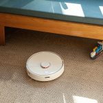 Ein Saugroboter f&auml;hrt auf einem Teppichboden auf eine Eckbank zu.