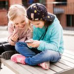 Zwei Kinder sitzen auf einer Bank im Freien und schauen gemeinsam auf ein Smartphone.