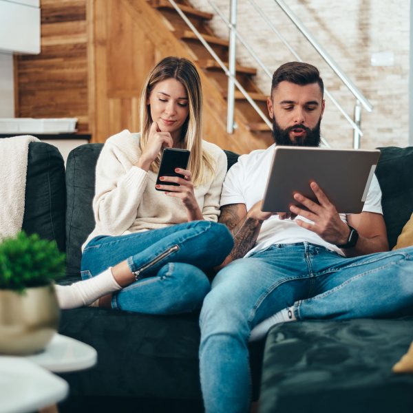Zwei Personen sitzen auf der Couch und haben jeweils ein iPhone und ein Tablet vor sich in der Hand.