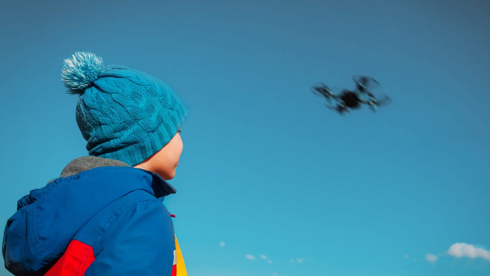 Ein Kid in Anorak und Pudelmütze blickt einer Drohne am Himmel hinterher.