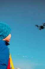 Ein Kid in Anorak und Pudelmütze blickt einer Drohne am Himmel hinterher.