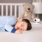Ein Kind liegt schlafend im Bett, im Hintergrund ein Teddy und ein Babyphone.