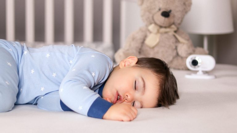 Ein Kind liegt schlafend im Bett, im Hintergrund ein Teddy und ein Babyphone.