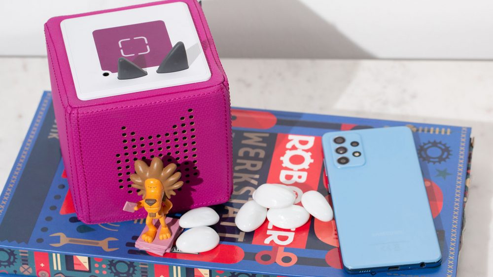Auf einem Tablett steht eine Toniebox neben einem Tonie, einem Smartphone und einigen weißen Steinen.