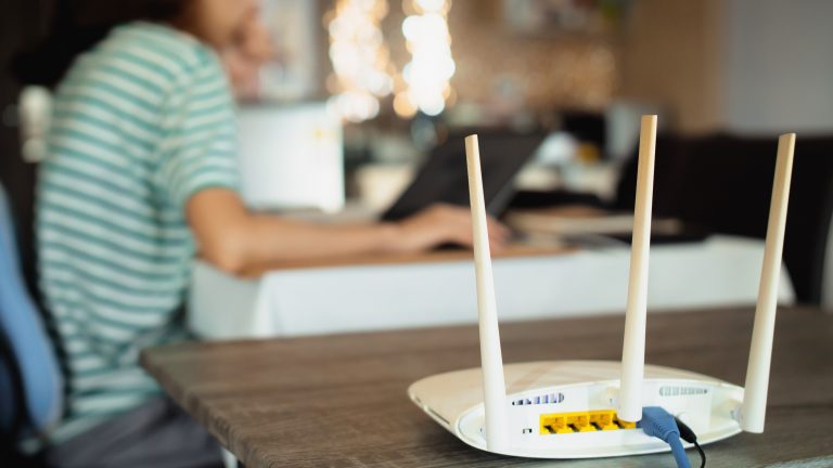 Ein WLAN-Router steht auf einem Tisch. Im Hintergrund ist eine Person an einem Laptop zu sehen.
