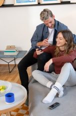 Fritzbox 7510 Ein Paar sitzt im Wohnzimmer auf einem Sofa, gemeinsam schauen sie auf ein Smartphone.