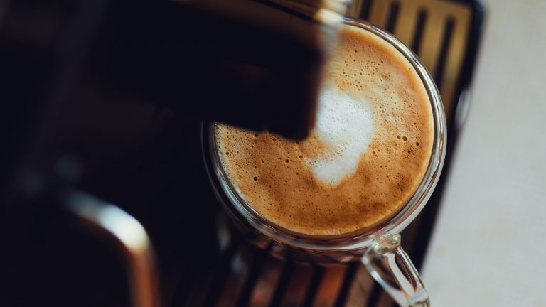 Unter dem Kaffeeauslauf eines Kaffeevollautomaten steht eine Kaffeetasse gefüllt mit Kaffee und Milchschaum.
