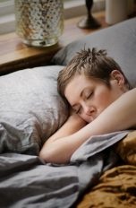 Eine Person liegt in einem Bett mit grauem Bezug und schläft, während eine Hand unter dem Kopf liegt.