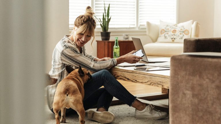 Eine Person sitzt auf dem Boden vor ihrem Wohnzimmertisch, darauf steht ein MacBook, in der Hand hält sie ein iPad, neben der Person steht ein Hund.