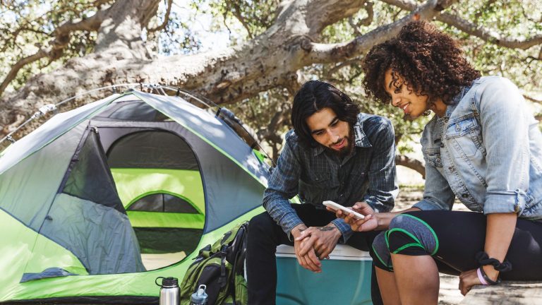 Zwei Personen sitzen im Freien vor einem Campingzelt und schauen gemeinsam auf ein Smartphone.