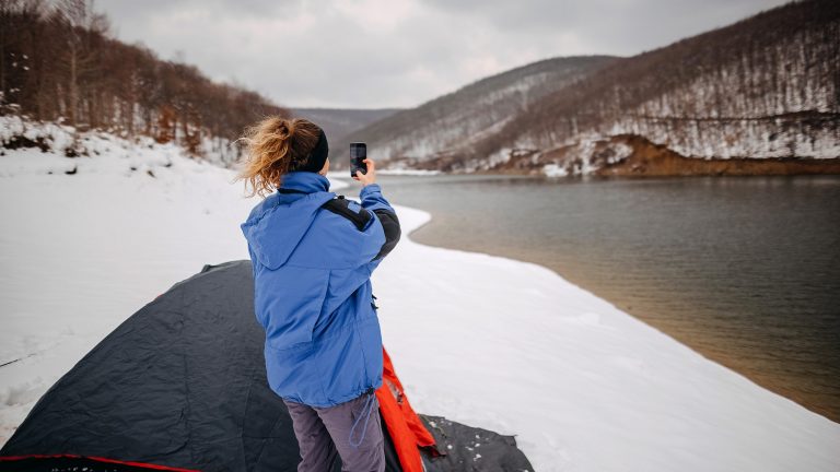 Eine Person steht vor einem Zelt im Schnee in der Nähe eines Flusses. In der Hand hält sie ein Smartphone, mit dem sie die Landschaft fotografiert.